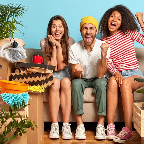 overjoyed three mixed race friends celebrate almost finishing repairing flat, pose on sofa, clench fists from joy. positive guy moves in new apartment, two female friends help him to unpack boxes